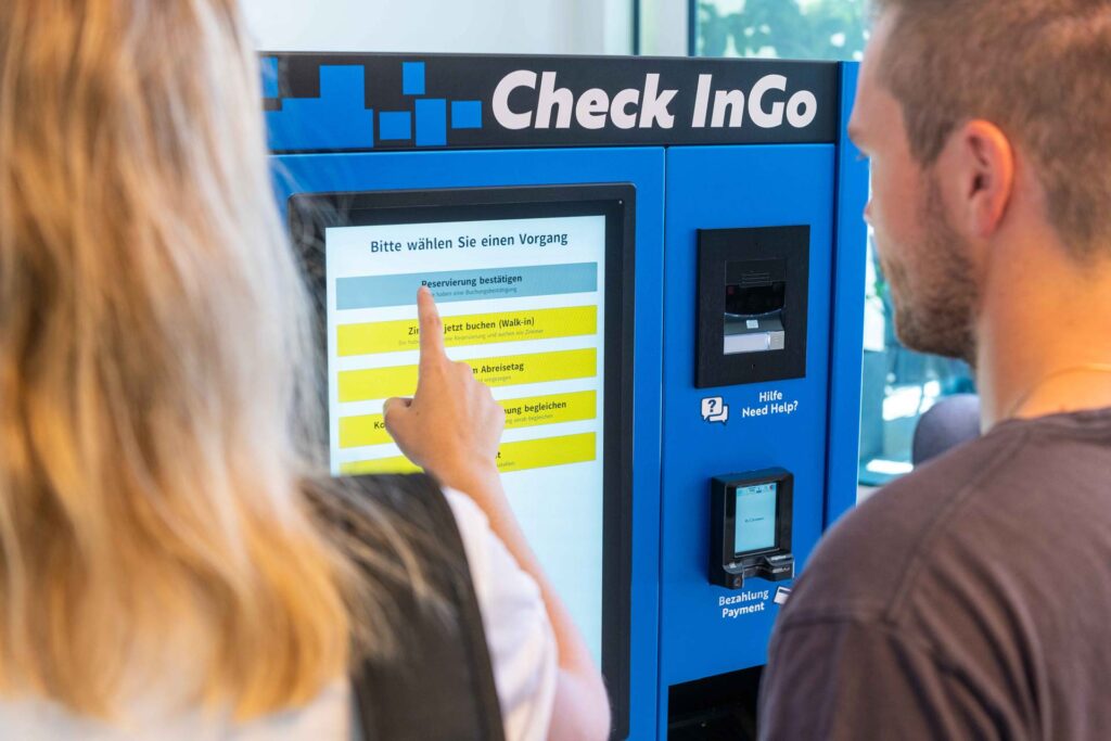 Mann und Frau wählen am CheckInGo Automaten von asut computer ihre Buchung per Touchscreen aus, Check-in und Walk-in Optionen verfügbar